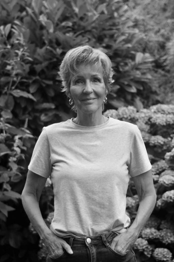 Smiling older woman with short hair wearing a light t-shirt and jeans standing outdoors with plants in the background